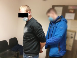 Zdjęcie przedstawia policjanta, który zakłada kajdanki zatrzymanemu mężczyźnie na ręce trzymane z tyłu. Obaj znajdują się w pomieszczeniu służbowym. Za policjantem widać drzwi oklejone kartkami z informacjami. Policjant jest ubrany w niebieską kurtkę, ma maseczkę ochronną na twarzy. Jest twarz jest rozmazana. Zatrzymany mężczyzna stoi do niego tyłem. Ma maseczkę ochronną na twarzy. Ubrany jest w czarną bluzę.