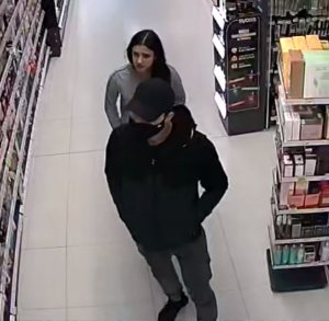Zdjęcie przedstawia mężczyznę ubranego na ciemno, który ma czapeczkę na głowie i czarną maseczkę na twarzy. anim stoi młoda kobieta z czarnymi długimi włosami i w szarej bluzie dresowej. W tle widać półki z asortymentem sklepowym.