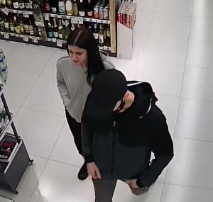 Zdjęcie przedstawia mężczyznę ubranego na ciemno, który ma czapeczkę na głowie i czarną maseczkę na twarzy. Za nim idzie młoda kobieta z czarnymi długimi włosami i w szarej bluzie dresowej. W tle widać półki z asortymentem sklepowym.