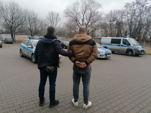 Zdjęcie przedstawia policjanta w ubraniu cywilnym, który przytrzymuje za lewe przedramię zatrzymanego mężczyznę. Mężczyzna ma kajdanki założone na ręce trzymane z tyłu. Mężczyzna jest ubrany w brązowo-czarną kurtkę, szare spodnie i białe buty sportowe. Policjant jest ubrany na ciemno. Obaj stoją na parkingu zewnętrznym. W tle widać zaparkowane oznakowane radiowozy.