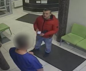 Zdjęcie przedstawia mężczyznę w czerwonej kurtce i niebieskich spodniach jeansowych, który stoi przed inną osobą ubraną w niebieski uniform. W tle widać wyposażenie pomieszczenia w postaci zielonych mebli.