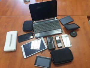 Zdjęcie przedstawia kilkanaście przedmiotów: laptop, tablet, nawigacje samochodowe, telefon komórkowy leżące na biurku.