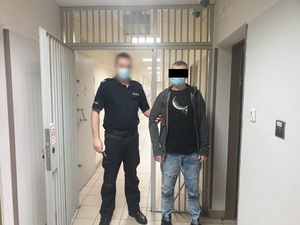Zdjęcie przedstawia policjanta w umundurowaniu służbowym, który przytrzymuje za prawe ramię zatrzymanego mężczyznę. Obaj stoją na wprost do obiektywu aparatu. Za nimi widać korytarz pomieszczenia dla osób zatrzymanych i okratowane drzwi. Policjant ma zasłoniętą twarz maseczką i górną połowę twarzy zasłania mu czarny prostokąt.
