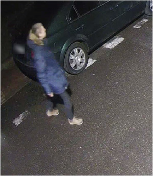 Zdjęcie przedstawia mężczyznę stojącego przy zaparkowanym samochodzie. Ubrany jest w granatową kurtkę z futrzanym kapturem, jeansowe spodnie i brązowe buty. Mężczyzna stoi tyłem do kamery monitoringu. Widać jego prawy profil.