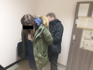 Zdjęcie przedstawia zatrzymaną kobietę, ma ona długie ciemne włosy. Ubrana jest w zieloną kurtkę zimową z kapturem. Jej twarz zasłania czarny prostokąt. Za nią stoi policjant w ubraniu cywilnym. Znajdują się oni w pomieszczeniu dla osób zatrzymanych.