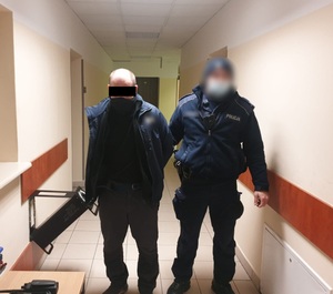 Zdjęcie przedstawia policjanta w granatowym mundurze, który przytrzymuje stojącego po jego prawej stronie zatrzymanego mężczyznę. Mężczyzna ten trzyma ręce z tyłu. Twarz zasłonięta ma czarną maseczką a okolice oczu czarnym prostokątem. Obaj stoją w korytarzu, na wprost obiektywu aparatu.