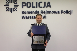 Zdjęcie przedstawia I Zastępcę Komendanta Rejonowego Policji Warszawa VI, który stoi w galowym mundurze, na tle jasnej ściany z napisem KOMENDA REJONOWA POLICJI WARSZAWA VI. Policjant trzyma w rękach tabliczkę pamiątkową.