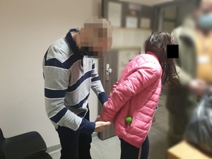 Zdjęcie przedstawia zatrzymaną kobietę. Ma ona długie ciemne włosy, jest ubrana w różową pikowaną kurtkę i ciemne spodnie. Trzyma ręce z tyłu. Za nią stoi policjant w ubraniu cywilnym, który zakłada jej kajdanki. Policjant ma bluzkę szarą w czarne szerokie pasy. Jego twarz jest rozmazana.