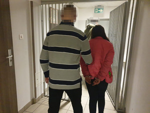 Zdjęcie przedstawia przedstawia zatrzymaną kobietę. Ma ona długie ciemne włosy, jest ubrana w różową pikowaną kurtkę i ciemne spodnie. Trzyma ręce z tyłu. Ma na nich założone kajdanki. Z jej lewej strony idzie policjant. Ma on na sobie cywilne ubranie - bluzkę szarą w czarne szerokie pasy. Oboje wychodzą z pomieszczenia dla osób zatrzymanych, po obu stronach widać pionowe pręty.