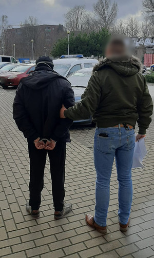 Zdjęcie przedstawia policjanta w ubraniu cywilnym, który stoi po prawej stronie zatrzymanego mężczyzny. Policjant ma na sobie ciemnozieloną kurtkę i niebieskie spodnie jeansowe. Zatrzymany mężczyzna jest ubrany na ciemno. Na rękach trzymanych z tyłu ma założone kajdanki. Obaj znajdują się na świeżym powietrzu. W tle widać zaparkowane pojazdy.