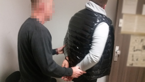 Zdjęcie przedstawia policjanta ubranego w cywilne ubrania, który stoi za zatrzymanym mężczyzną. Ma on kajdanki na rękach trzymanych z tyłu.