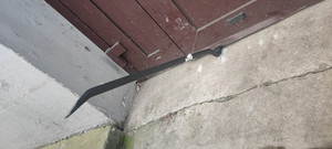 Zdjęcie przedstawia czarny łom wsunięty pod drzwi wejściowe koloru brązowego