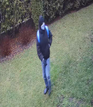 Zdjęcie przedstawia mężczyznę znajdującego się na podwórzu. Jest on w wieku około 25-30 lat, szczupłej budowy ciała, ma czarną kurtkę z kapturem, czarną czapkę, czarne rękawiczki, jeansowe spodnie, przez ramiona ma przewieszone niebieskie paski plecaka, na twarzy ma założony tzw. komin lub chustę.