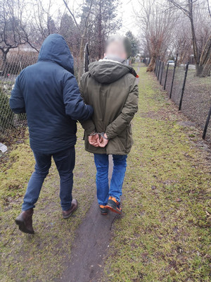 Zdjęcie przedstawia policjanta w ubraniu cywilnym, który prowadzi zatrzymanego mężczyznę. Mężczyzna ten ma kajdanki założone na rękach trzymanych z tyłu. Jest ubrany w zimową kurtkę w kolorze ciemnozielonym i granatowe spodnie jeansowe.