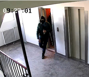 Zdjęcie przedstawia mężczyznę wychodzące z windy na klatkę schodową. Jest on w wieku około 30-35 lat, ubrany w zieloną kurtkę, czarną bluzę z kapturem, granatowe spodnie, brązowe buty, na twarzy ma założoną czarną maseczkę.