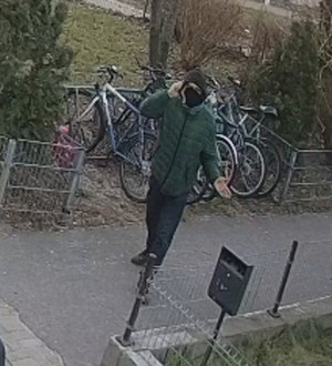 Zdjęcie przedstawia mężczyznę stojącego na dworze przy stojaku z rowerami. Jest on w wieku około 30-35 lat, ubrany w zieloną kurtkę, czarną bluzę z kapturem, granatowe spodnie, brązowe buty, na twarzy ma założoną czarną maseczkę.