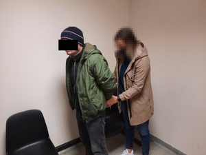 Zdjęcie przedstawia policjantkę w pomieszczeniu dla osób zatrzymanych, która zakłada kajdanki na ręce trzymane z tyłu zatrzymanemu mężczyźnie. Mężczyzna ten ma na sobie zieloną kurtkę, granatowe spodnie jeansowe i zimową czapkę. Twarz ma zasłoniętą czarnym prostokątem.