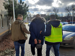 Zdjęcie przedstawia policjantów prowadzących mężczyznę po dziedzińcu. Mężczyzna ten ma kajdanki na rękach założonych z tyłu. Jeden z policjantów ma odblaskową kamizelkę. W tle widać zapakowane pojazdy.