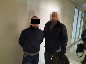 Zdjęcie przedstawia policjanta w ubraniu cywilnym, który prowadzi zatrzymanego mężczyznę. Twarz mężczyzny zasłania czarny prostokąt. Jest on ubrany na ciemno.