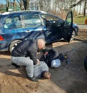 Zdjęcie przedstawia policjanta w ubraniu cywilnym, który klęka przy zatrzymanych leżących na ziemi mężczyznach. Obok nich jest zaparkowany niebieski pojazd, który ma otwarte prawe przednie drzwi od pasażera.