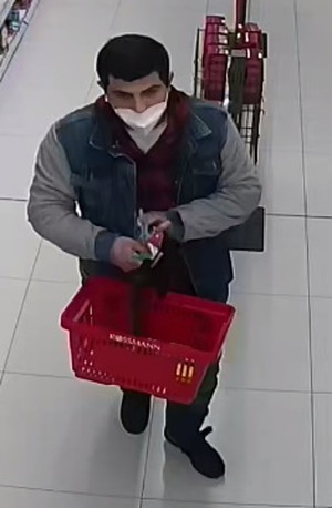 Zdjęcie przedstawia mężczyznę w wieku około 30 lat, około 170 cm wzrostu, czarne krótkie włosy, ubrany w czerwono - czarną bluzę z kapturem z widocznym wzorem w kratę, niebieską kurtkę z szarymi rękawami, niebieskie spodnie, czarne buty, w prawym uchu ma białą słuchawkę bezprzewodową. Dolną część twarzy zasłania mu jasna maseczka ochronna. Mężczyzna niesie przed sobą, trzymając w obu rękach czerwony koszyk sklepowy.