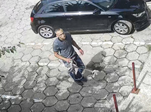 Zdjęcie przedstawia mężczyznę w wieku około 40-45, normalnej budowy ciała, ma siwe włosy, czesane na bok, ubrany jest w spodnie jeansowe koloru niebieskiego, czarne buty sportowe, niebieską koszulkę z krótkim rękawem z granatowym kołnierzykiem oraz czarną kurtkę. W tle widać zaparkowany czarny samochód.
