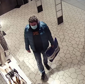 Zdjęcie przedstawia mężczyznę wewnątrz sklepu. Ma on ciemną karnację, czarne włosy, ubrany jest w niebieską kurtkę pikowaną, czarną bluzę, spodnie typu jeans, sportowe obuwie, twarz zasłania mu maseczka ochronna, w ręku trzyma reklamówkę z logiem „Intersport”. Jest skierowany na wprost kamery.
