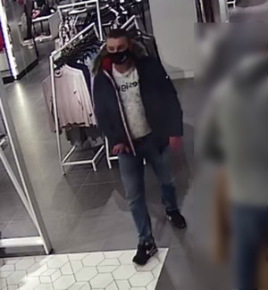 Zdjęcie przedstawia mężczyznę wewnątrz sklepu odzieżowego. Jest on w wieku około 30 lat, ubrany w spodnie typu jeans, jasną bluzę, granatową kurtkę z kapturem obszytym futrem, ciemne obuwie sportowe, na twarzy ma czarną maseczkę ochronną. W tle widać wieszaki z odzieżą.