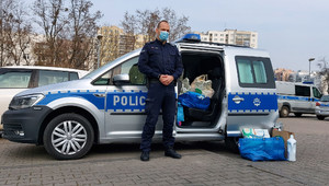 Zdjęcie przedstawia umundurowanego policjanta, który stoi przy policyjnym oznakowanym radiowozie. Obok samochodu stoją torby z kupionymi artykułami. W tle widać inne zaparkowane pojazdy.
