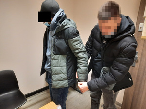 Zdjęcie przedstawia policjanta ubranego w cywilne ubrania, który zakłada kajdanki na ręce trzymane z tyłu zatrzymanemu mężczyźnie. Mężczyzna ten ma czarną czapkę i kolorową kurtkę. Twarz policjanta jest zasłonięta.