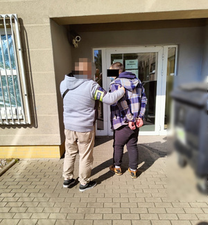 Zdjęcie przedstawia policjanta ubranego w cywilne ubrania, który prowadzi zatrzymanego mężczyznę. Ma on kajdanki na rękach trzymanych z tyłu. Stoją przodem do budynku.
