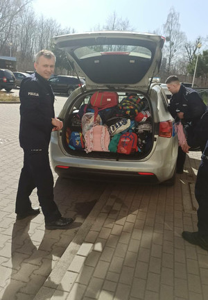 Zdjęcie przedstawia umundurowanych policjantów, którzy stoją przy radiowozie, klapa bagażnika jest otwarta, w środku widać poukładane kolorowe plecaki.