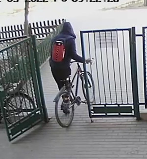 Zdjęcie przedstawia osobę wyprowadzającą rower z posesji. Ubrana jest w niebieską kurtkę z kapturem, czarne spodnie dresowe,  niebieskie buty sportowe, posiada czerwony plecak.