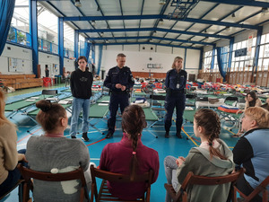 Zdjęcie przedstawia halę sportową. Przed siedzącymi na krzesłach tyłem do aparatu osobami stoi policjant i policjantka w granatowych mundurach i kobieta w czarnej bluzie i spodniach jeansowych. W tle widać zielono-srebrne leżaki.