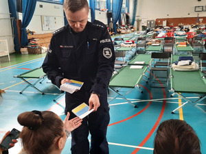 Zdjęcie przedstawia halę sportową. Przed siedzącymi na krzesłach tyłem do aparatu osobami stoi policjant w granatowym mundurze, który podaje żółto-niebieską ulotkę jednej osobie. W tle widać zielono-srebrne leżaki.