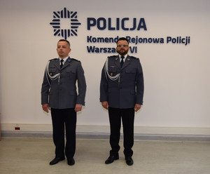 Zdjęcie przedstawia policjantów w galowych mundurach, którzy stoją na sali na tle ściany z napisem POLICJA KOMENDA REJONOWA POLICJI WARSZAWA VI.