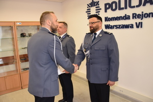 Zdjęcie przedstawia trzech policjantów w galowych mundurach, którzy stoją na sali. Jeden z nich podaje rękę wyróżnionemu policjantowi.