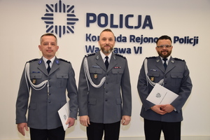 Zdjęcie przedstawia trzech policjantów w galowych mundurach, którzy stoją na sali na tle ściany z napisem POLICJA KOMENDA REJONOWA POLICJI WARSZAWA VI. Dwaj z nich trzymają w dłoniach białe teczki.