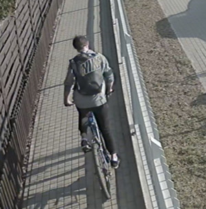 Zdjęcie przedstawia jadącego rowerem po chodniku mężczyznę. Mężczyzna jest ubrany w szarą bluzę z kapturem, czarne spodnie, sportowe buty, plecak z napisem „NIKE”. Jest skierowany frontem do obiektywu kamery.