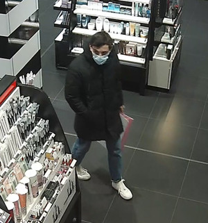Zdjęcie przedstawia mężczyznę wewnątrz sklepu, stoi on przy regale z kosmetykami. Jest on w wieku około 30-35 lat, ma około 175 cm wzrostu, czarne włosy, jest ubrany w czarną kurtkę do kolan z kapturem, niebieskie spodnie, białe sportowe buty; w ręku trzyma torbę prezentową z czarnym n/n napisem, na twarzy ma założoną maseczkę ochronną zakrywającą usta i nos.