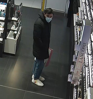 Zdjęcie przedstawia mężczyznę wewnątrz sklepu, stoi on przed regałem z kosmetykami. Jest on w wieku około 30-35 lat, ma około 175 cm wzrostu, czarne włosy, jest ubrany w czarną kurtkę do kolan z kapturem, niebieskie spodnie, białe sportowe buty; w ręku trzymał torbę prezentową z czarnym n/n napisem, na twarzy ma założoną maseczkę ochronną zakrywającą usta i nos.