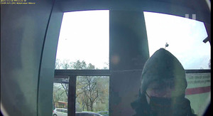 Zdjęcie przedstawia osobę w czapce i maseczce na twarzy, która stoi w oszklonym pomieszczeniu. Znajduje się na wprost kamery.