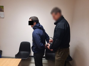 Zdjęcie przedstawia policjanta w pomieszczeniu dla osób zatrzymanych, który zakłada kajdanki zatrzymanego mężczyźnie na ręce trzymane z tyłu. Policjant jest ubrany na ciemno, jego twarz i głowa są zasłonięta filtrem komputerowym. Zatrzymany mężczyzna ma ciemne włosy, jest ubrany w granatową bluzę z kapturem. Obaj stoją lewym profilem do obiektywu aparatu.