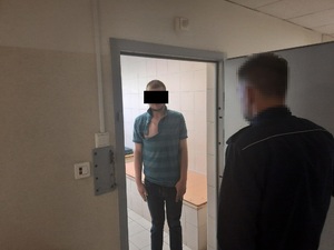 Zdjęcie przedstawia policjanta w granatowym mundurze, który stoi przy otwartych drzwiach pomieszczenia dla osób zatrzymanych, w środku przodem do obiektywu aparatu stoi zatrzymany młody mężczyzna. Ma on koszulkę na krótki rękaw, twarz zasłania mu czarny prostokąt.