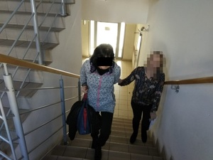 Zdjęcie przedstawia policjantkę w ubraniu cywilnym, która wprowadza po schodach zatrzymaną kobietę. Policjantka ma twarz zasłoniętą komputerowym filtrem. Kobieta ma czarne rozpuszczone włosy, część twarzy zasłania jej czarny prostokąt. Obie wchodzą po schodach, znajdują się na wprost obiektywu aparatu.