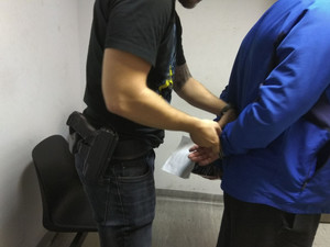 Zdjęcie przedstawia dwie sylwetki mężczyzn. Jedna jest ubrana w niebieską kurtkę, trzyma ręce z tyłu. Druga osoba zakłada kajdanki na ręce trzymane z tyłu.