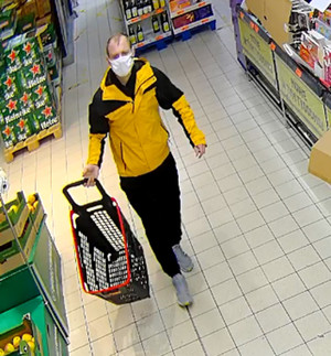 Zdjęcie przedstawia mężczyznę idącego przez sklep z czarnym pustym wózkiem sklepowym. Mężczyzna jest w wieku ok 35 lat, ubrany jest w czarne spodnie, obuwie sportowe w kolorze białym, kurtkę w kolorze żółto-czarnym, na twarzy ma maseczkę ochronną.