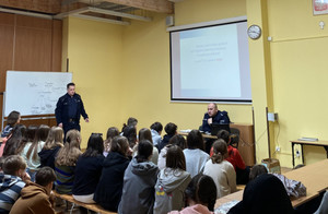 Zdjęcie przedstawia dwóch policjantów w granatowych mundurach, którzy stoją przed uczniami zgromadzonymi w sali. Uczniowie siedzą na krzesłach. Są tyłem do obiektywu aparatu.