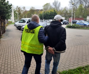 Zdjęcie przedstawia policjanta prowadzącego dziedzińcem zatrzymanego mężczyznę. policjant ma na sobie seledynową odblaskową kamizelkę z napisem POLICJA. Twarz policjanta jest zasłonięta komputerowym filtrem. Mężczyzna ma założone kajdanki na rękach trzymanych z tyłu. Ubrany jest w ciemną kurtkę i jasną czapeczkę z daszkiem. Obaj stoją tyłem do obiektywu aparatu. W tle widać zaparkowane pojazdy osobowe i radiowozy oznakowane.