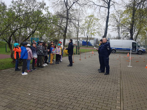 Zdjęcie przedstawia policjantów w granatowych mundurach, którzy stoją przed uczniami na dziedzińcu.